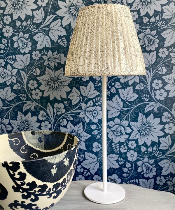 A vintage silk lampshade on a white candlestick base on a table with black and white bowl and floral folk wallpaper in dark blue behind.