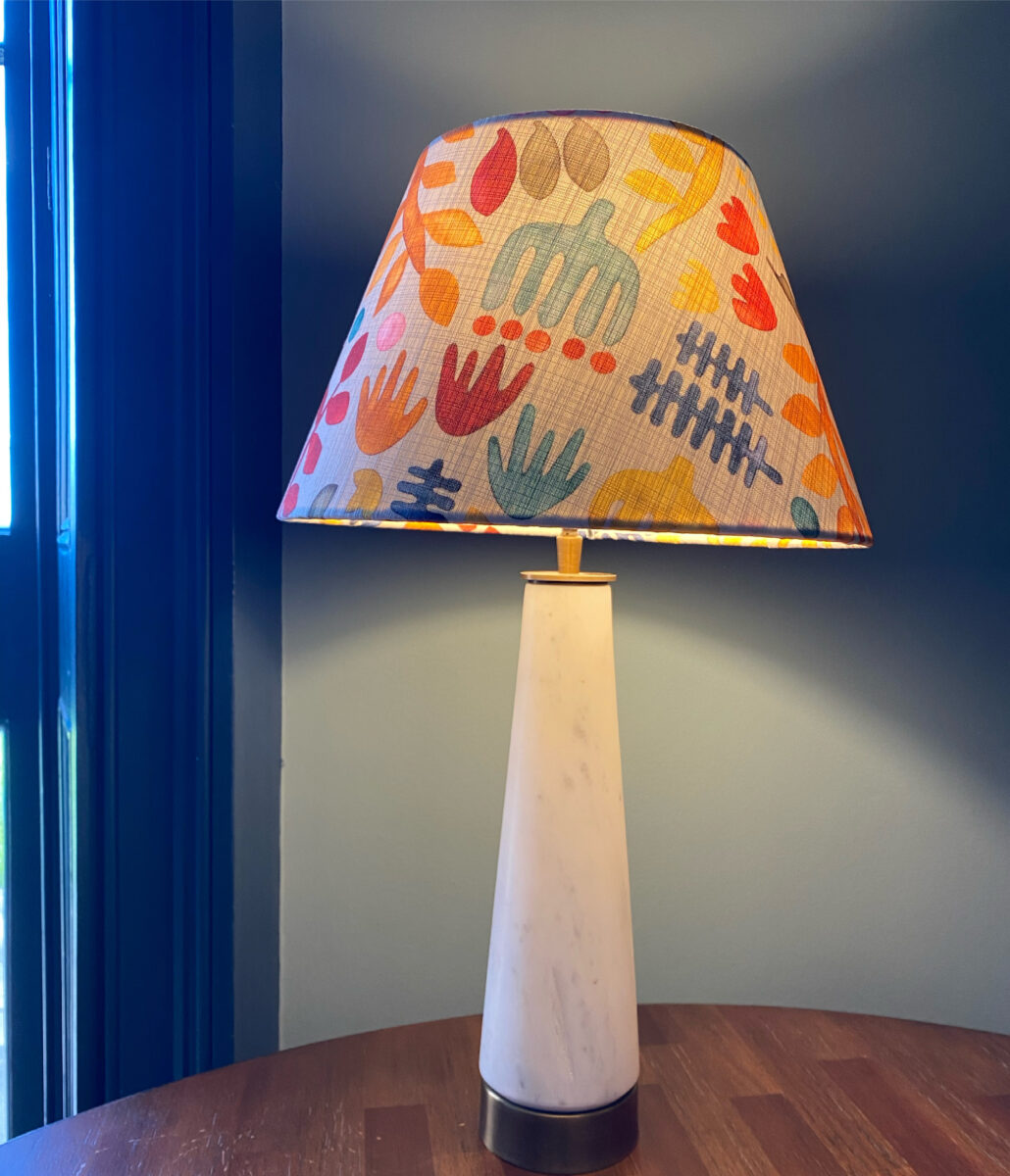 A colourful, West African-inspired tribal print coolie lampshade on a white conical lamp base against a blue backdrop.