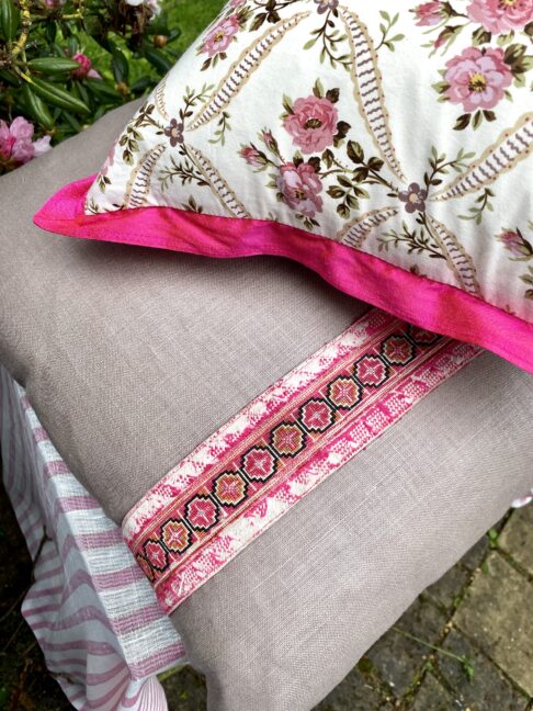 A vintage floral fabric cushion with pink trim on top of glazed taupe linen cushions with an embroidery trim.