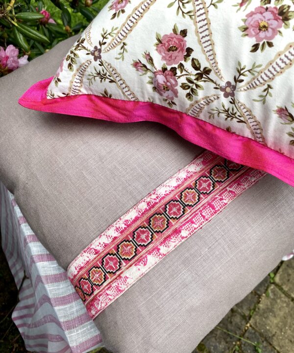 A vintage floral fabric cushion with pink trim on top of glazed taupe linen cushions with an embroidery trim.