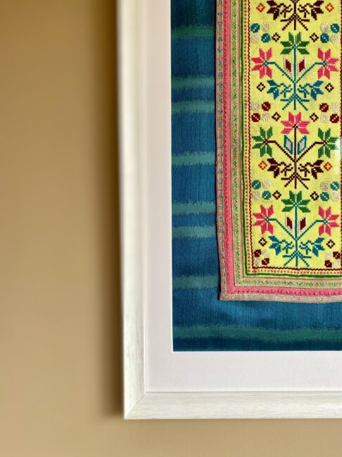 Bright acid yellow embroidery art textile framed in a white frame against teal silk.