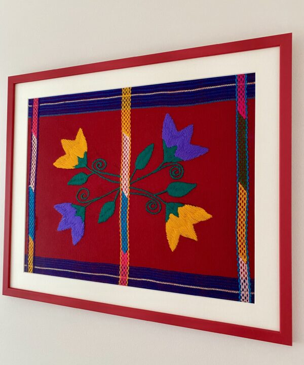 Shot from an angle a colourful, framed red art embroidery textile from Mexico.