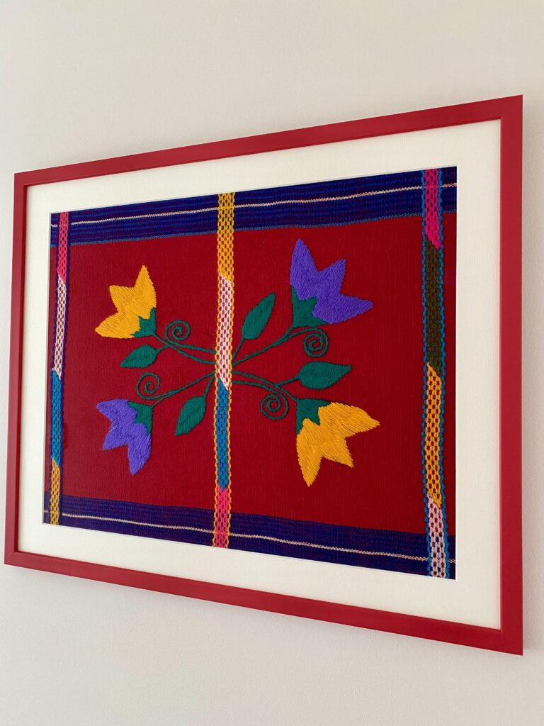 Shot from an angle a colourful, framed red art embroidery textile from Mexico.