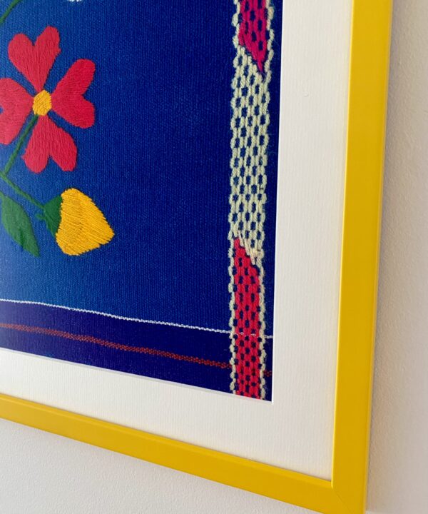 Corner detail of a yellow frame housing an embroidered Mexican textile in blue and pink.