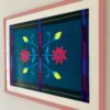 A teal Mexican embroidered textile in a pale pink frame shot from an angle.