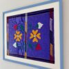 A Mexican, lavender/violet-hued framed embroidery art textile in pale blue shown from an angle.