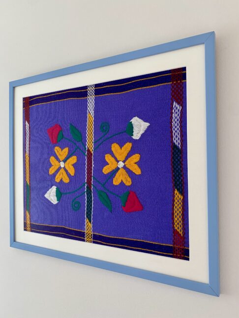 A Mexican, lavender/violet-hued framed embroidery art textile in pale blue shown from an angle.