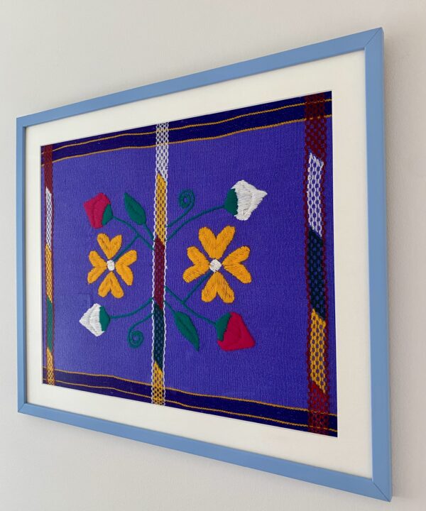 A Mexican, lavender/violet-hued framed embroidery art textile in pale blue shown from an angle.