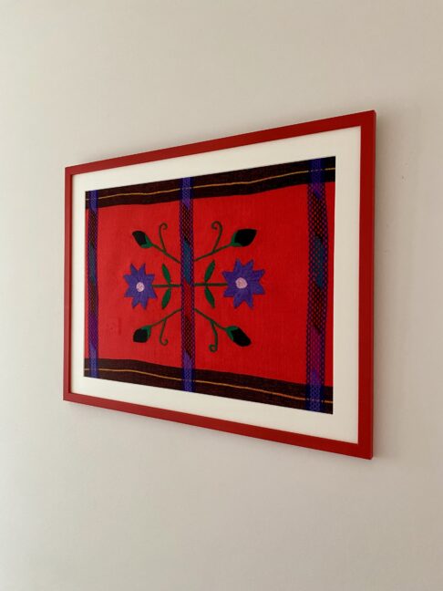 A framed floral embroidery art textile from Mexico in red and coral shown shot from the side.