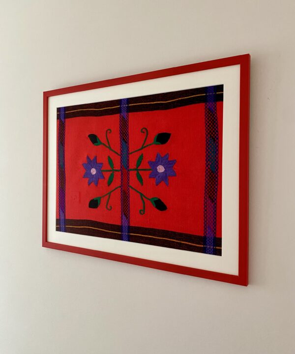 A framed floral embroidery art textile from Mexico in red and coral shown shot from the side.