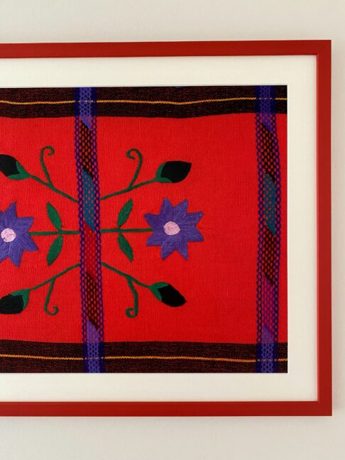 Violet embroidered flowers and buds on a bright coral ethnic Mexican cloth framed with a red picture frame.