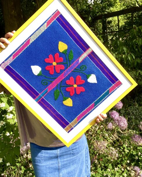 Bright yellow framed Mexican textile art depicting floral embroidery.