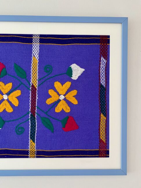 A Mexican embroidery textile in violet, yellow and pink in a baby blue frame.