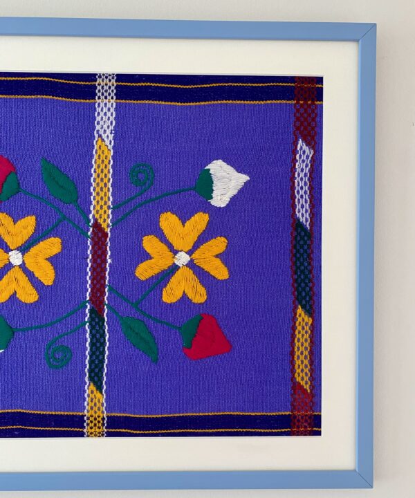 A Mexican embroidery textile in violet, yellow and pink in a baby blue frame.