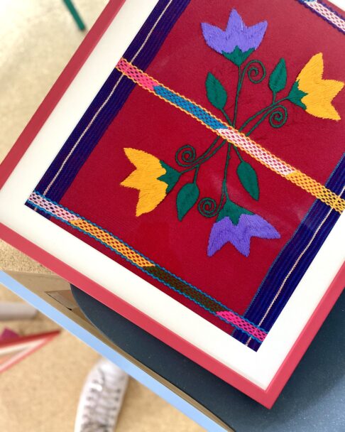 Embroidered, floral Mexican wall art in red, yellow and blue with a red frame.