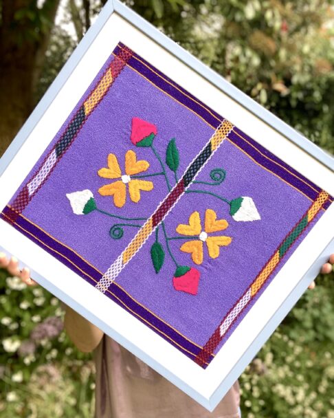 A baby blue framed Mexican textile in lilac, yellow and pink held in a garden setting.