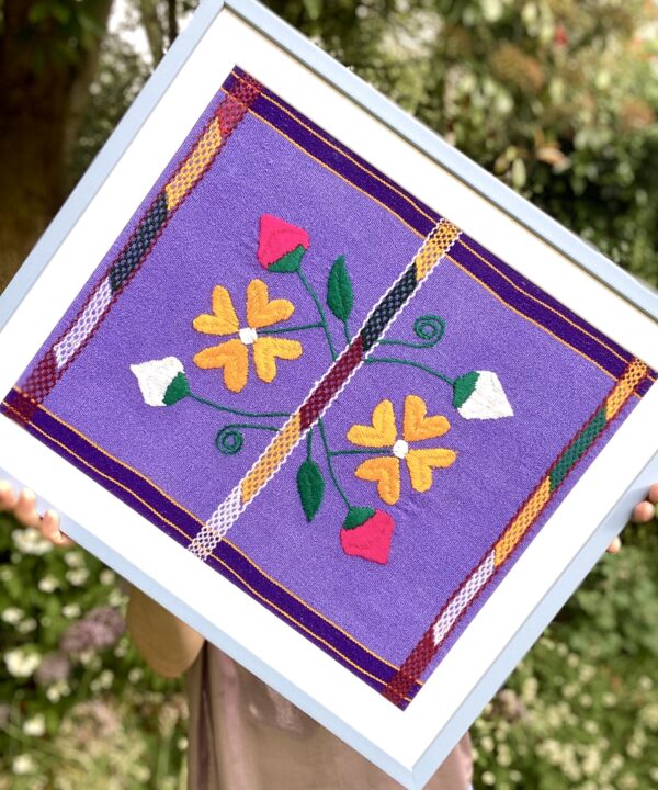 A baby blue framed Mexican textile in lilac, yellow and pink held in a garden setting.