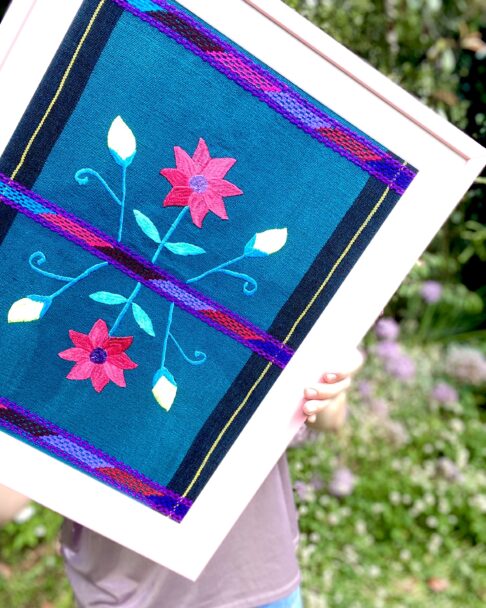 A turquoise and pink framed textile from Mexico in a baby pink frame in an outdoor setting.