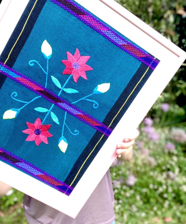 A turquoise and pink framed textile from Mexico in a baby pink frame in an outdoor setting.