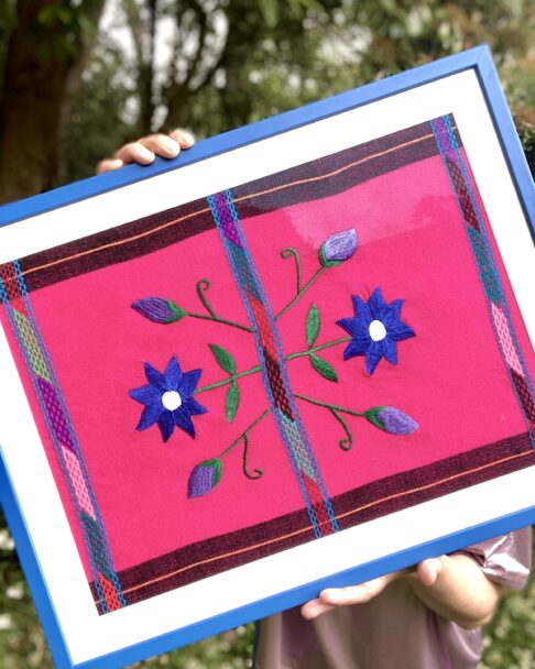 A Royal blue framed Mexican textile in blue and hot pink held in a garden setting.