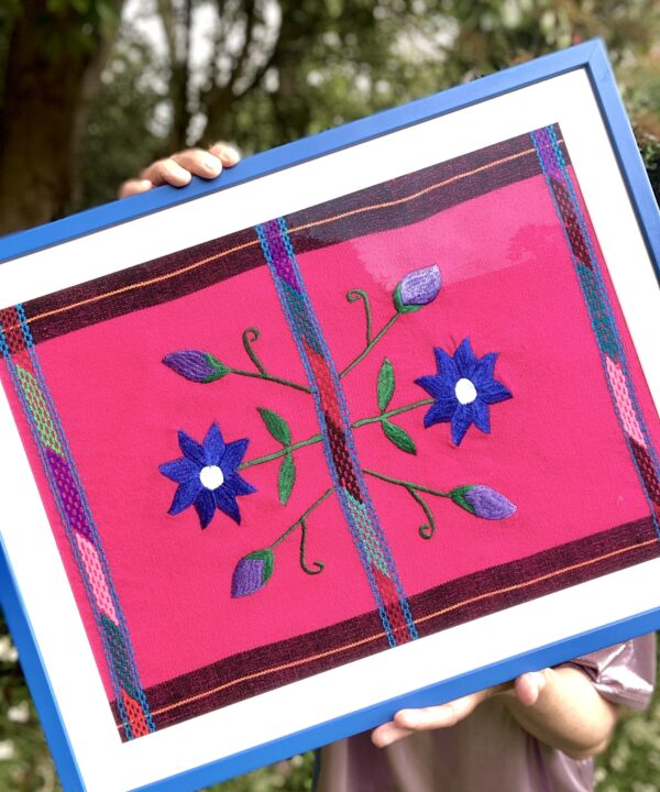 A Royal blue framed Mexican textile in blue and hot pink held in a garden setting.