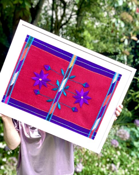 Purple Mexican embroidered flower art framed in pink in a garden setting.