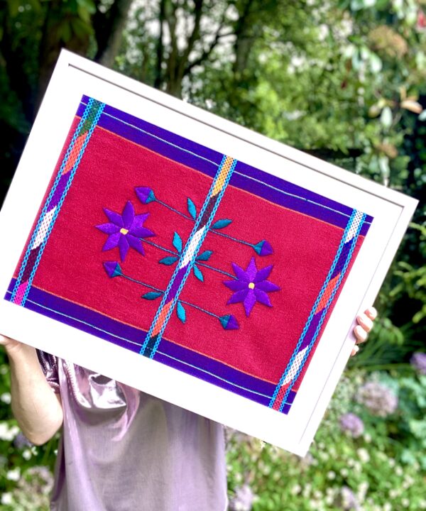 Purple Mexican embroidered flower art framed in pink in a garden setting.