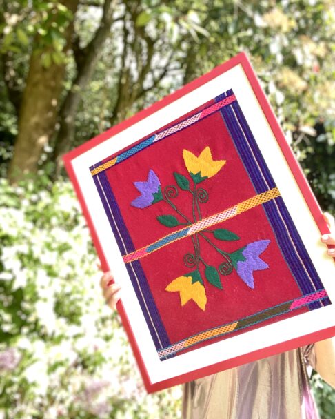 A red-framed Mexican textile being held in a garden setting.