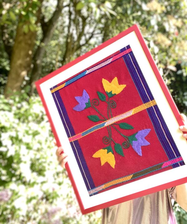 A red-framed Mexican textile being held in a garden setting.