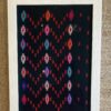 A framed textile showcasing colourful embroidery on a black cotton background, typical of South East Asian patterning.