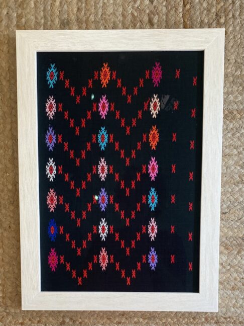 A framed textile showcasing colourful embroidery on a black cotton background, typical of South East Asian patterning.