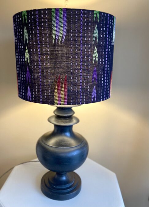 An illuminated, large black table lampshade with colourful embroidered details showing light diffusion and styled here with a pedestal lamp base from Pooky Lights.