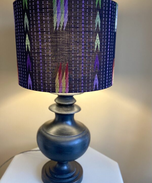 An illuminated, large black table lampshade with colourful embroidered details showing light diffusion and styled here with a pedestal lamp base from Pooky Lights.