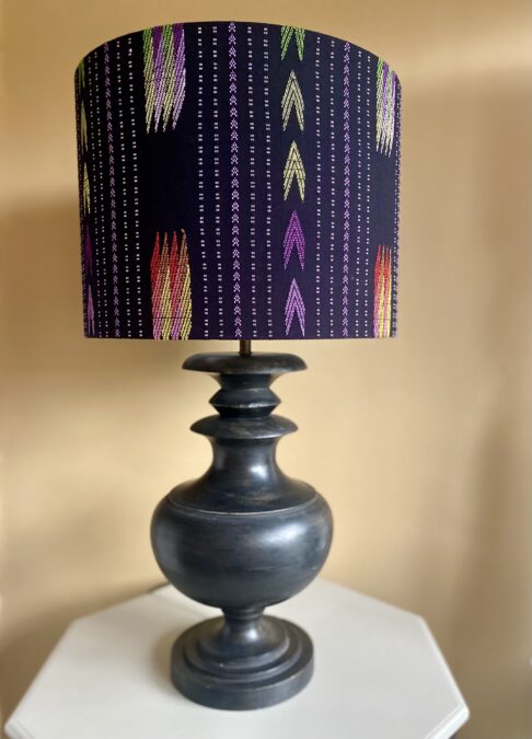 A large black cotton table lampshade in a drum shape with embroidered Southeast Asian motifs styled here on a wooden urn-shaped base from Pooky LIghts.