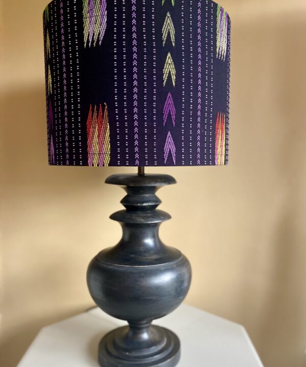 A large black cotton table lampshade in a drum shape with embroidered Southeast Asian motifs styled here on a wooden urn-shaped base from Pooky LIghts.