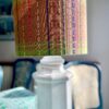 A patterned green lampshade in a drum design handmade from Southeast Asian cotton fabric with a colourful, embroidered stitch design, shown here on a white ceramic lamp base on a dining room sideboard.