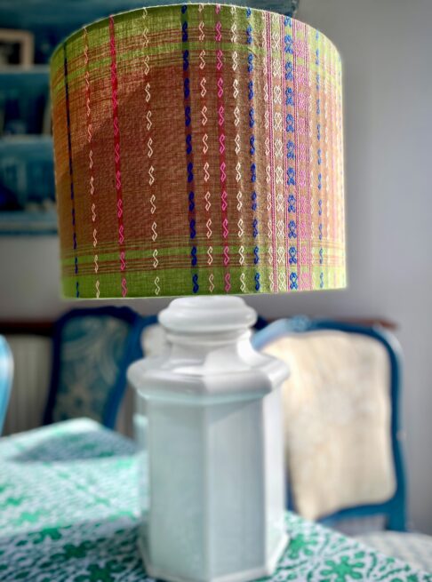 A patterned green lampshade in a drum design handmade from Southeast Asian cotton fabric with a colourful, embroidered stitch design, shown here on a white ceramic lamp base on a dining room sideboard.