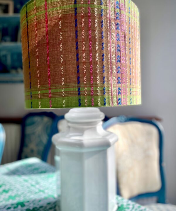 A patterned green lampshade in a drum design handmade from Southeast Asian cotton fabric with a colourful, embroidered stitch design, shown here on a white ceramic lamp base on a dining room sideboard.