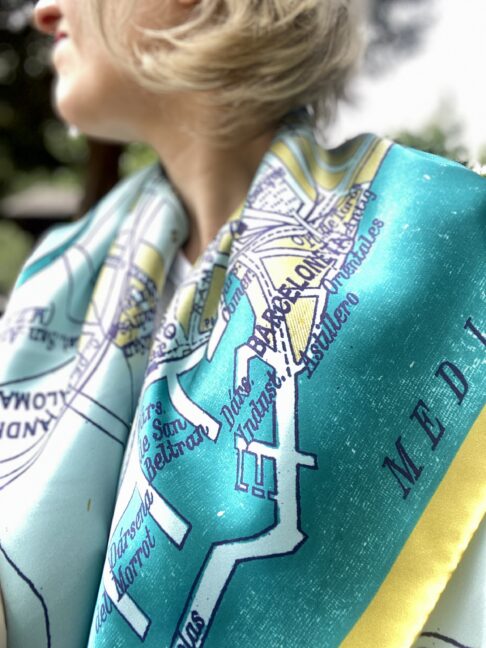 Model wearing a luxury silk scarf in teal and gold with a vintage map of Barcelona printed on it.