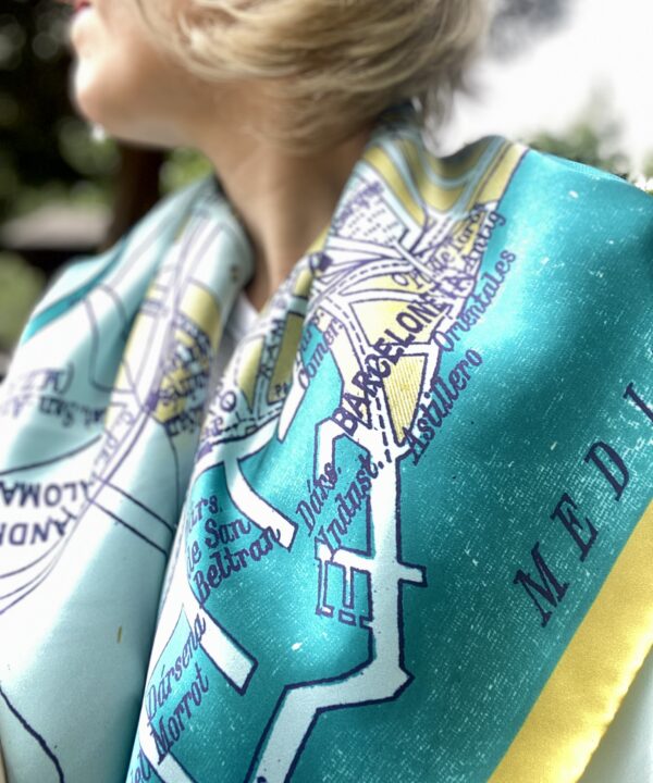 Model wearing a luxury silk scarf in teal and gold with a vintage map of Barcelona printed on it.