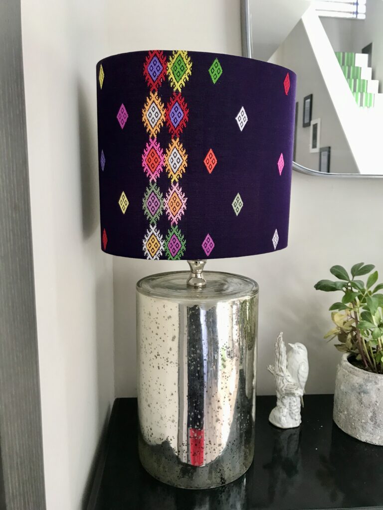 A colourful Thai textile drum lampshade on a silvered lamp base on a hall console.