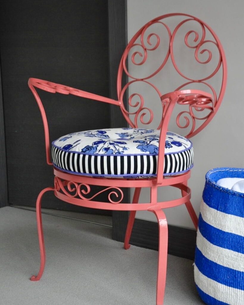 A coral-pink, French, wrought iron chair dating from the 1930s with a blue and white striped and floral cushion and a stripy basket.