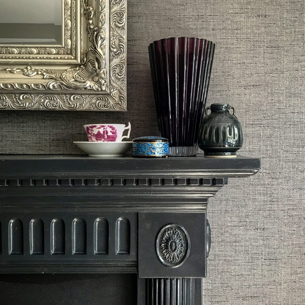 A gloss grey painted mantlepiece with various decorative accessories displayed against mottled grey wallpaper with a silver mirror above.