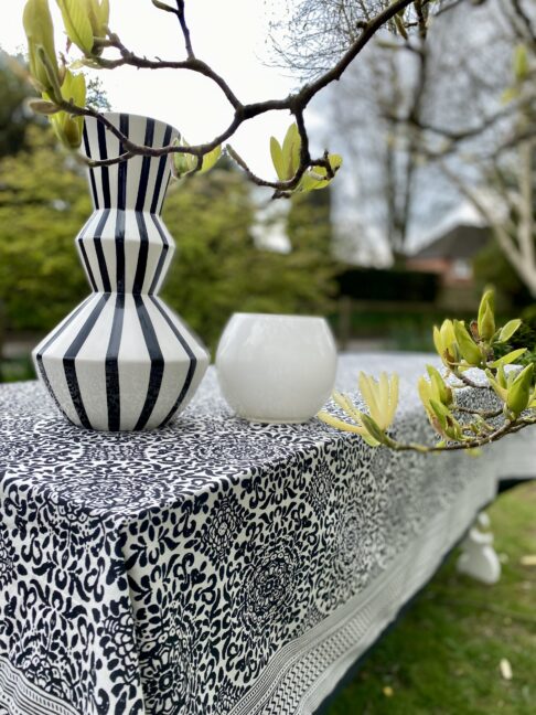 A stripy black and white vase sits on a Mughal-style, block-printed tablecloth in black and white, in a garden setting.