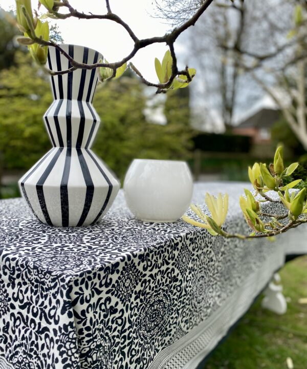 A stripy black and white vase sits on a Mughal-style, block-printed tablecloth in black and white, in a garden setting.