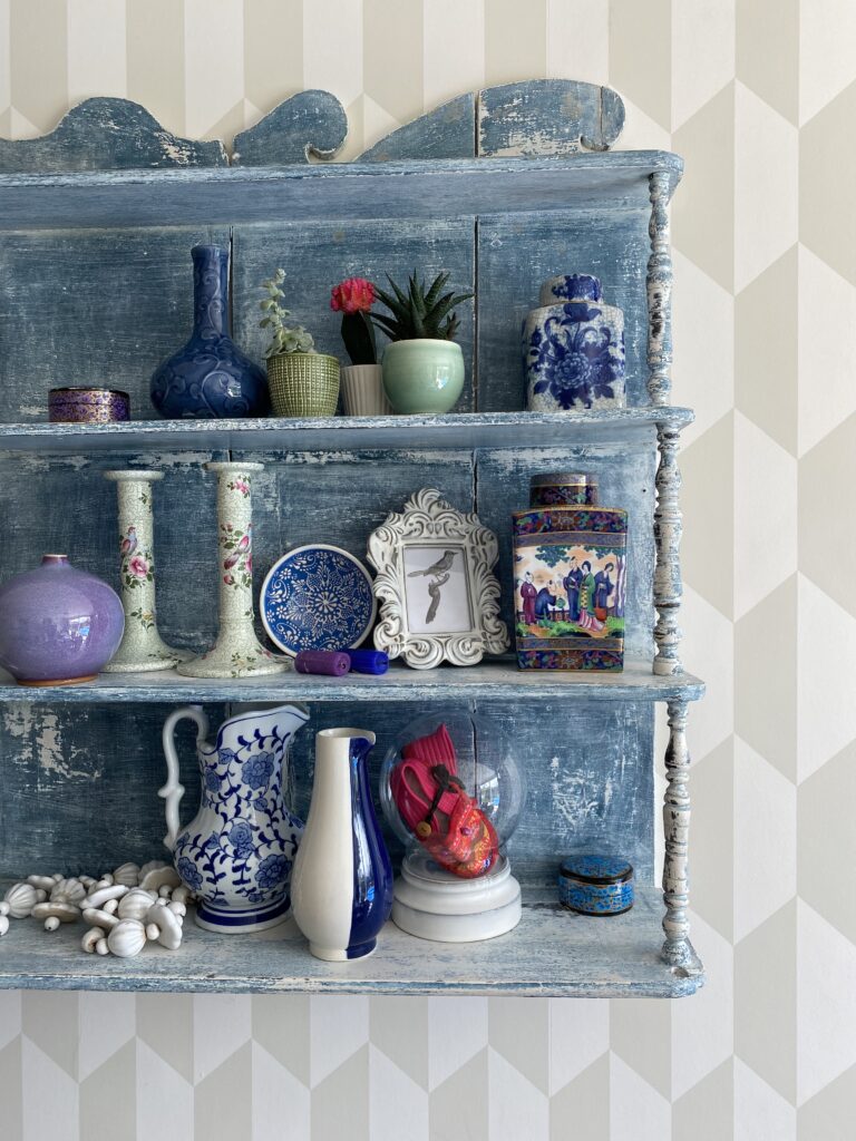 A distressed, vintage, Swedish wall shelf in pale blue against muted geometric wallpaper. The shelves are file with ecorative ceramics, knick-knacks and collected finds.