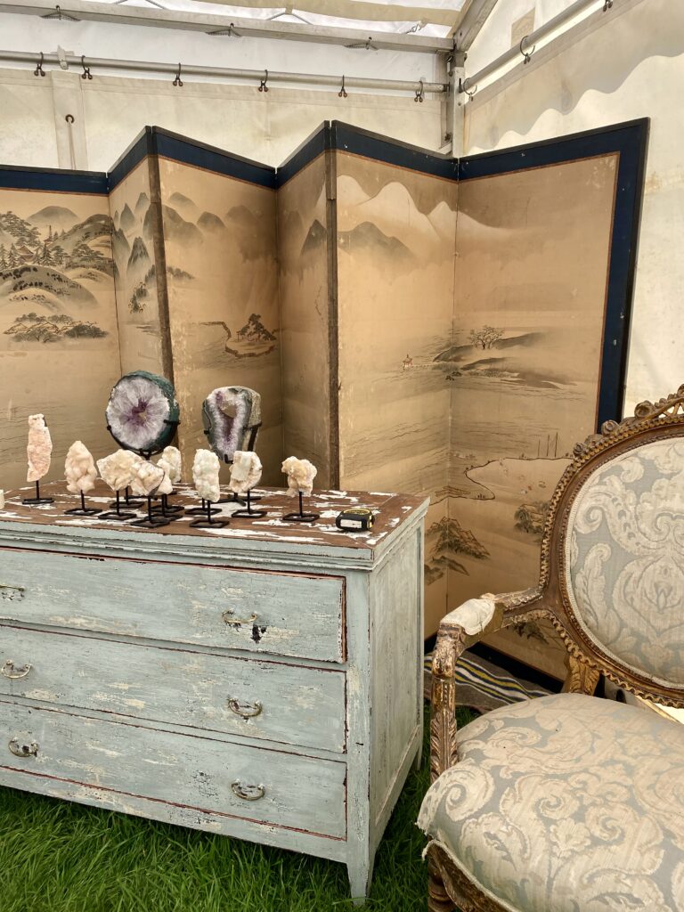 A large, folding screen depicting an Asian landscape in an antiques fair tent next to a vintage chest of drawers and gilt-framed, French chair.