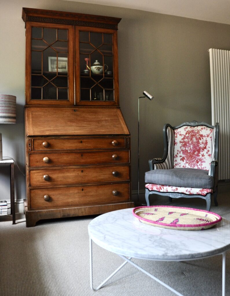 A tall, wooden bureau dresser with glazed bookshelves sits in a contemporary sitting room with a French chair, modern lighting and marble coffee table.