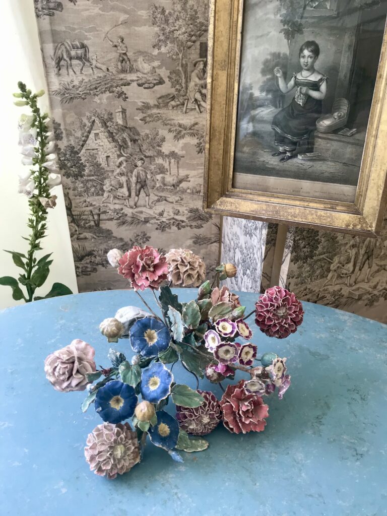 A bunch of antique, decorative ceramic flowers in a pretty palette of blues and pinks on a blue surface in front of vintage prints and wallpaper.