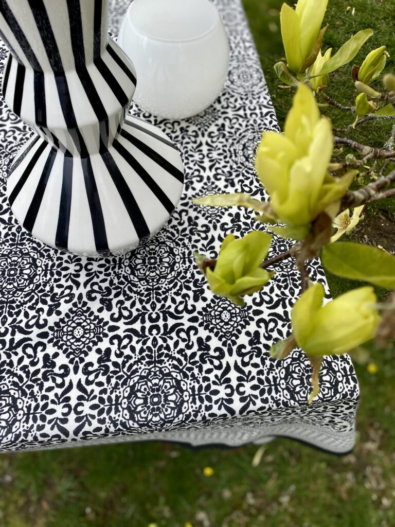 An overhead shot of a monochrome block print tablecloth with stripy black and white vase and yellow magnolia in the foreground.